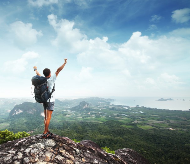 Young man with backpack standing with raised hands on top of a mountain and enjoying valley view